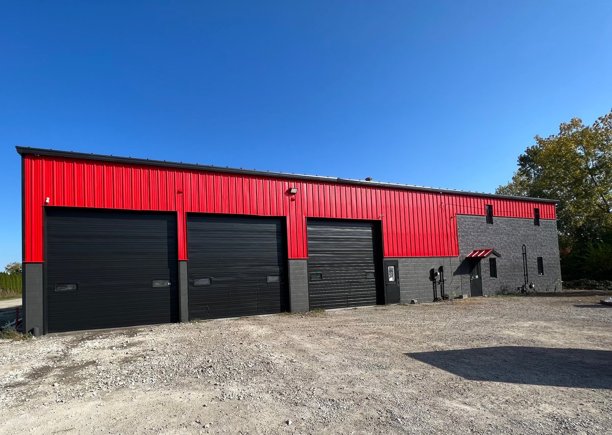 Modern red metal warehouse building with three black garage doors and gravel driveway under clear blue sky