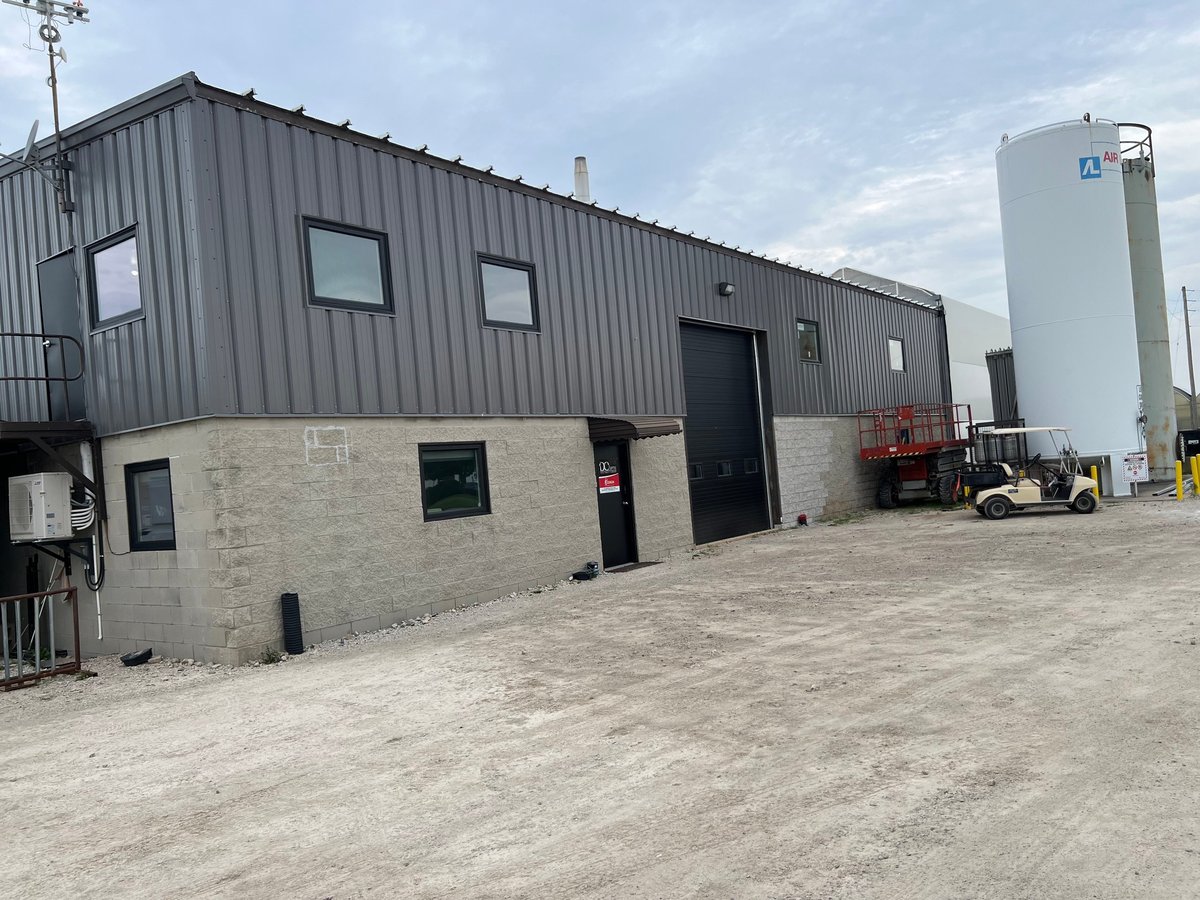 Industrial warehouse building with metal and concrete facade, black doors, and adjacent storage tank on gravel yard