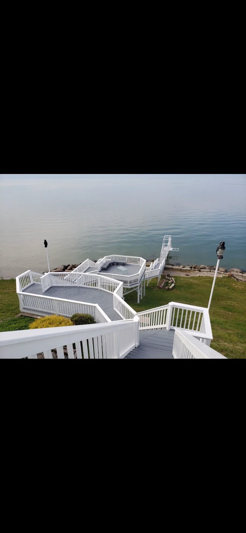 White wooden deck with railings overlooking calm waterfront and grassy lawn, viewed from above